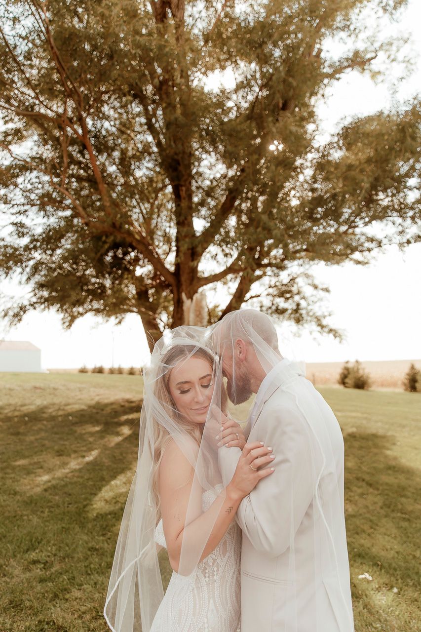 A tender wedding moment between a couple standing under a large tree at golden hour in an open field.