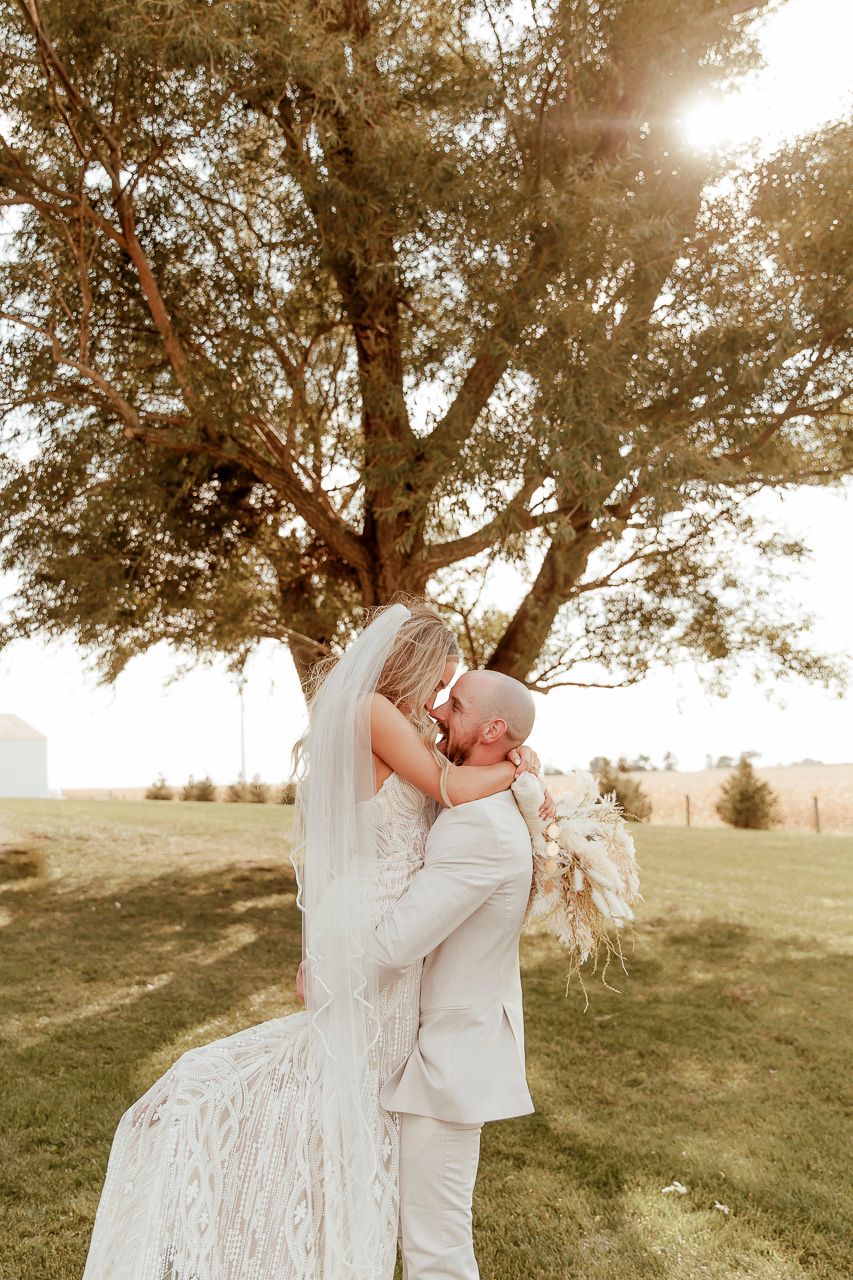 A loving couple shares a romantic moment under a sunlit tree during their outdoor wedding ceremony.