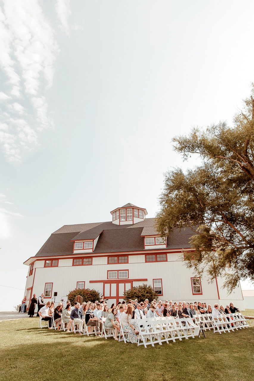 An outdoor wedding ceremony setup with white chairs on a lawn in front of a large red and white barn-style building.
