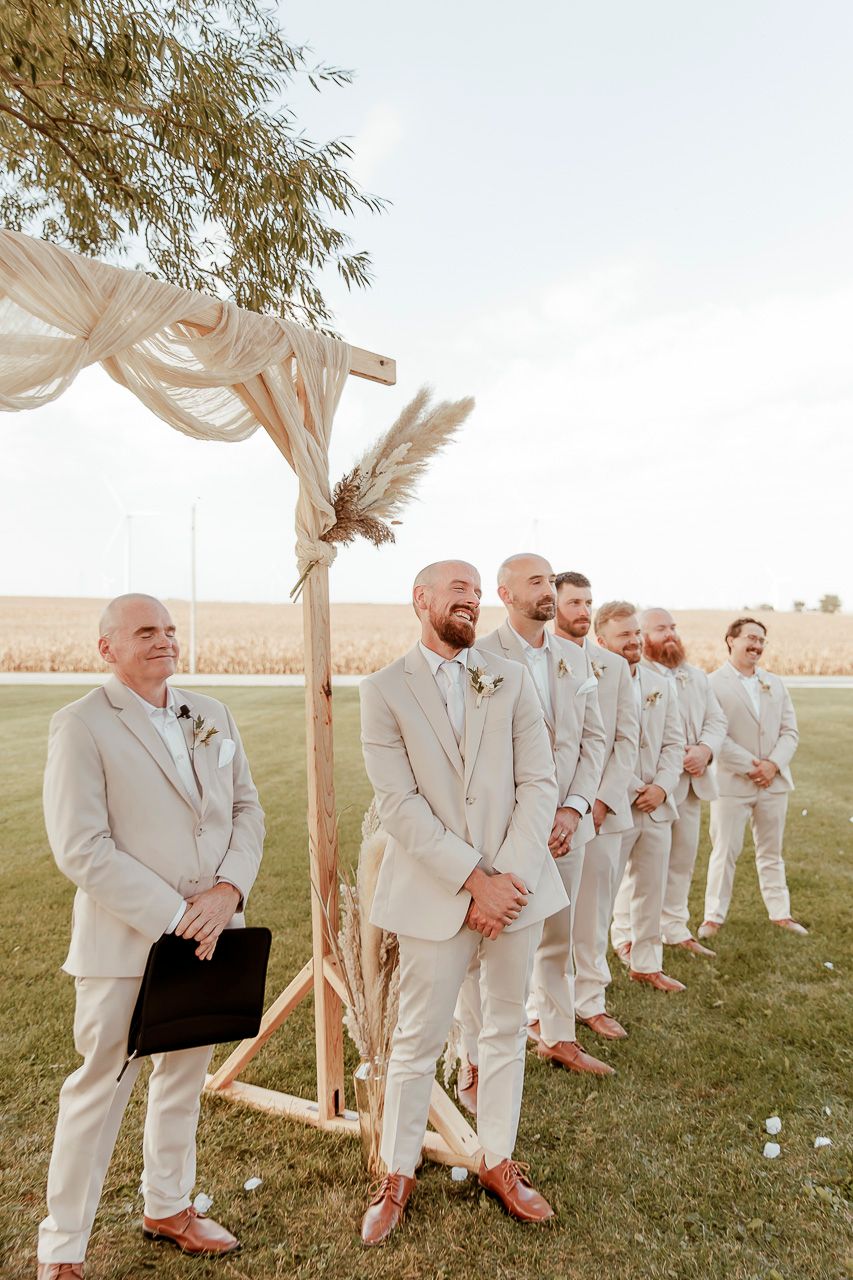 Groomsmen stand in line wearing light beige suits under a fabric-draped wooden arch at an outdoor wedding ceremony.