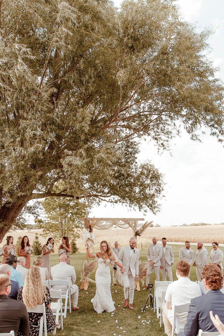 An outdoor summer wedding ceremony takes place under a large shady tree with white chairs set up on grassy lawn.