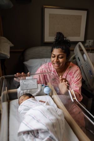 A patient in a pink robe joyfully records a special moment with their newborn in a hospital bassinet.
