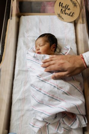 A sleeping newborn baby swaddled in a striped white blanket lies in a wooden bassinet as a hand gently touches them.