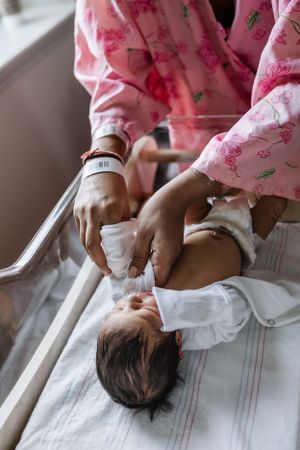 A newborn baby wearing white clothing lies on hospital bedding while being cared for by someone in pink floral scrubs.