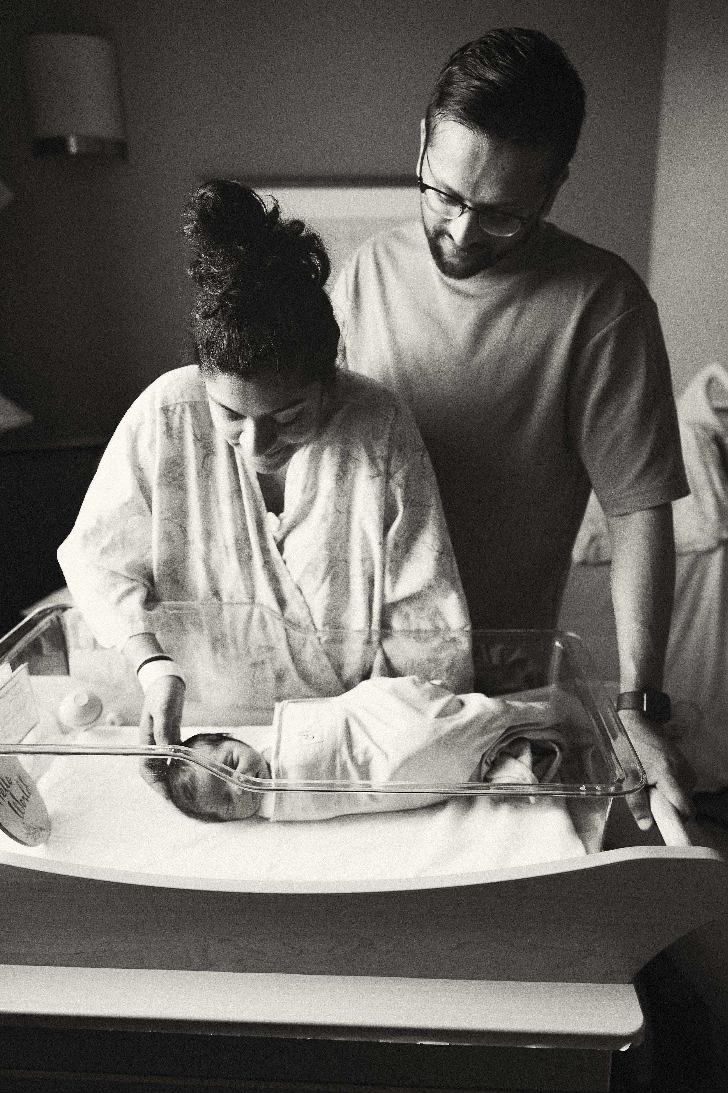 Black and white photo of parents looking at their newborn baby in a hospital bassinet.