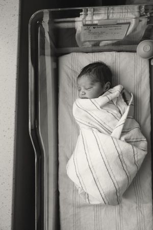 A newborn baby swaddled in a striped white blanket sleeps peacefully in a hospital bassinet.