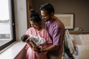 A loving couple stands near a window while tenderly holding their newborn baby wrapped in a blanket.