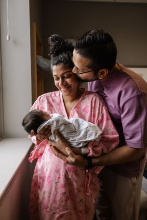 Two parents share a tender moment while holding their newborn baby by a window in a hospital room.