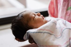 A peaceful sleeping newborn baby wearing a white patterned onesie rests in soft lighting.