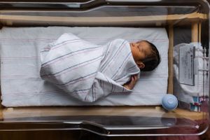 Baby swaddled in white striped blanket sleeps peacefully in hospital bassinet with medical equipment nearby.