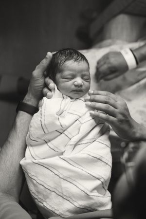 A newborn baby swaddled in white blankets is cradled by caring hands in this tender black and white photo.