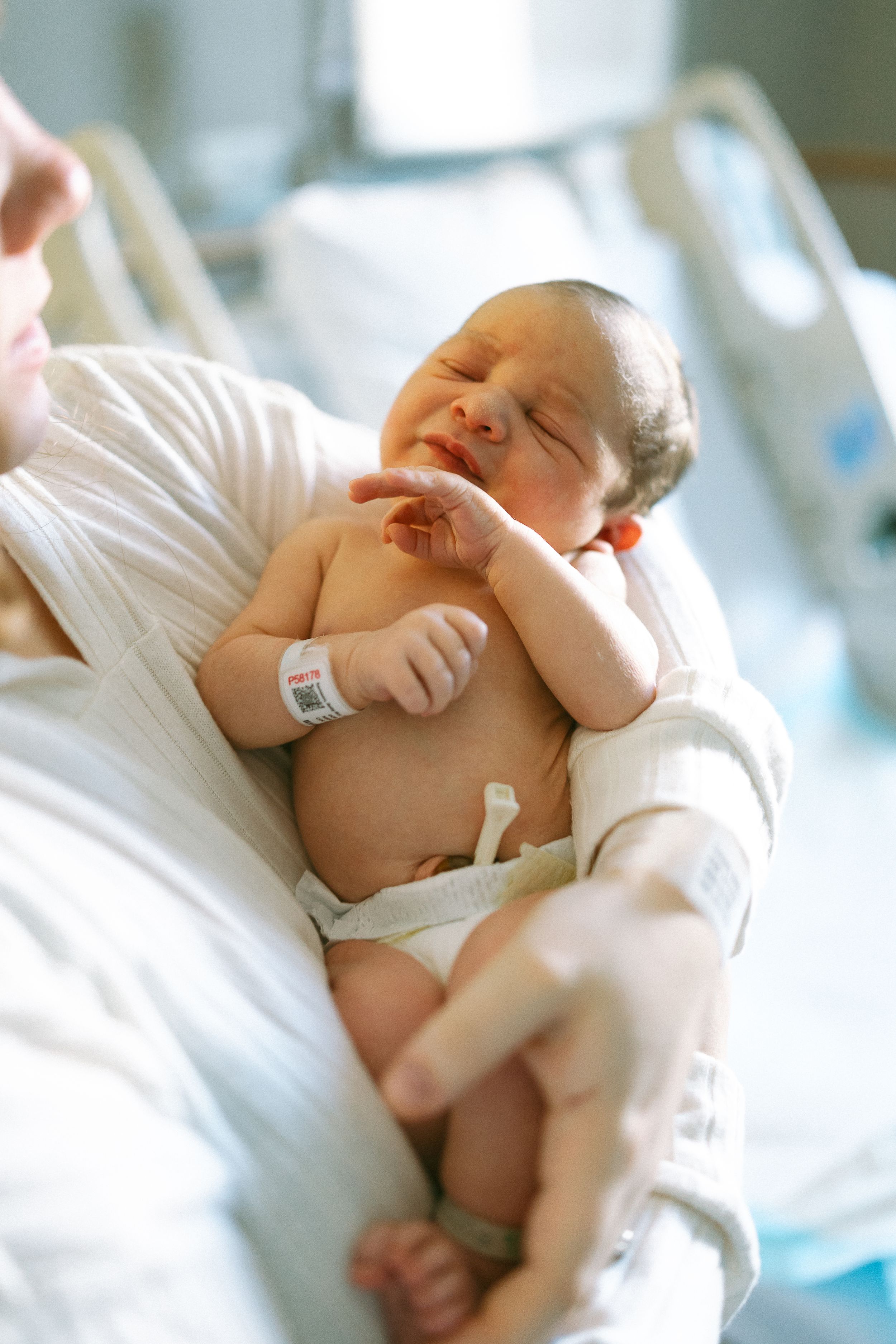 A newborn baby sleeps peacefully in white hospital bedding while wearing a medical bracelet.
