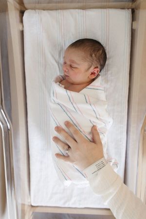 Newborn baby wrapped in striped white blanket resting in hospital bassinet while caregiver's hand gently touches.