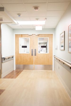 Double wooden doors with windows at the end of a clean hospital hallway with handrails and ceiling mounted lighting.