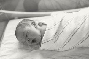 Black and white photo of a newborn baby swaddled in white blanket laying on hospital bed.