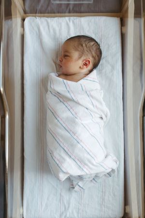 A sleeping newborn swaddled in a striped white blanket lies peacefully in a bassinet.