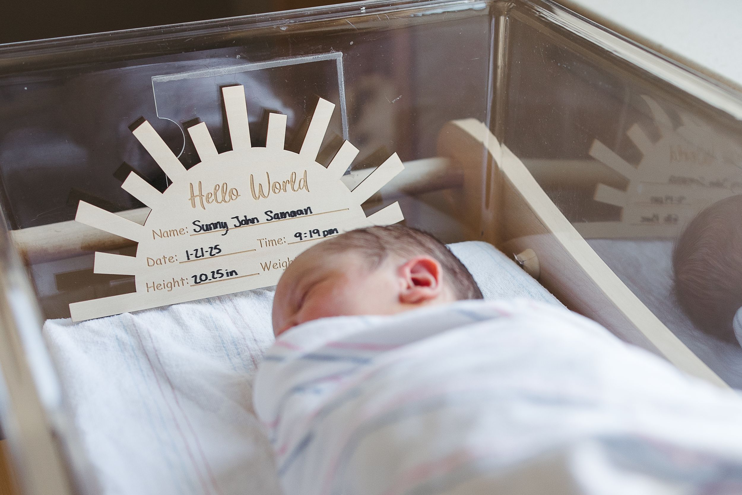 A newborn sleeping in a hospital bassinet with a decorative name tag displaying birth information above.