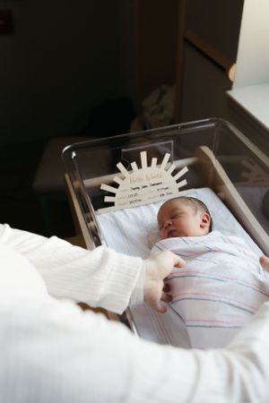 A newborn baby sleeps peacefully in a hospital bassinet while being swaddled in white blankets.