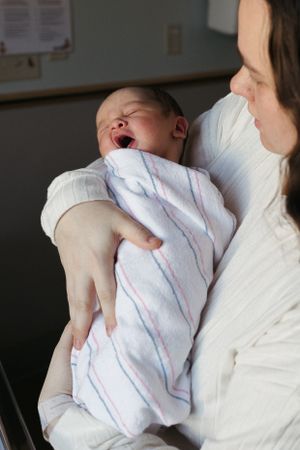 A newborn baby yawns while being held in white blanket against a caregiver wearing white.