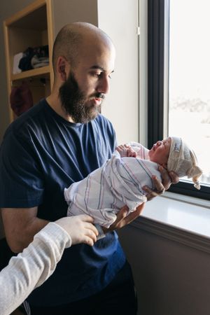 A bearded man in a navy t-shirt tenderly holds a swaddled newborn baby while standing by a window.