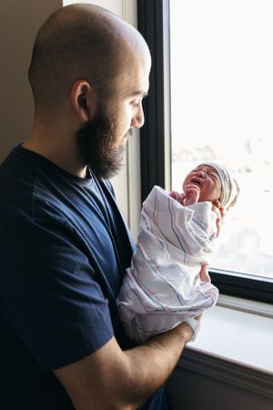 A tender moment by the window as a newborn baby in white swaddle is held while crying.