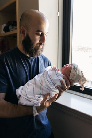 A bearded person in a blue shirt holds a newborn baby wrapped in a striped blanket near a window.