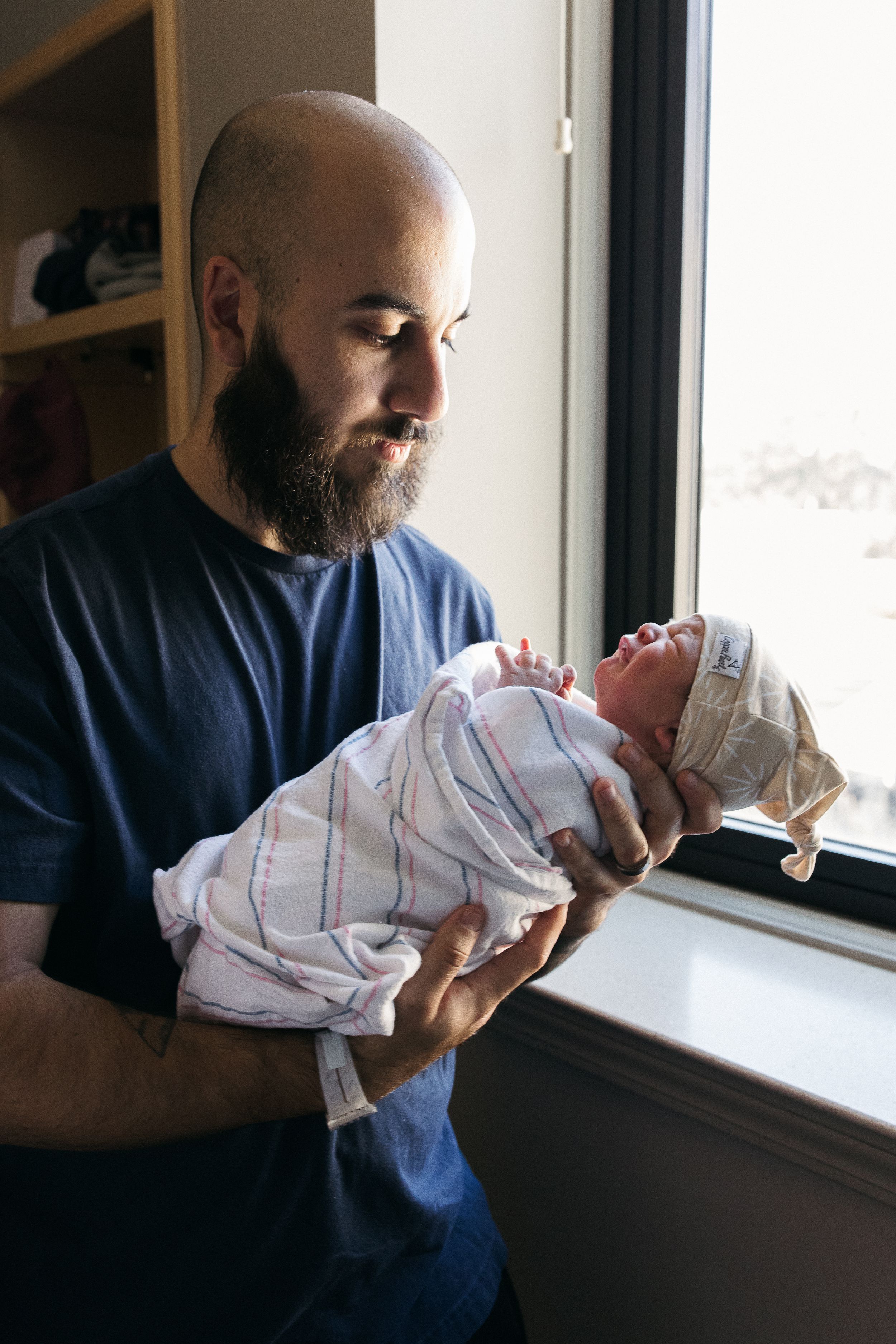 A bearded person in a blue shirt holds a newborn baby wrapped in a striped blanket near a window.