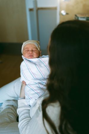 A swaddled sleeping newborn baby wrapped in striped blanket rests on hospital bed while being watched over.