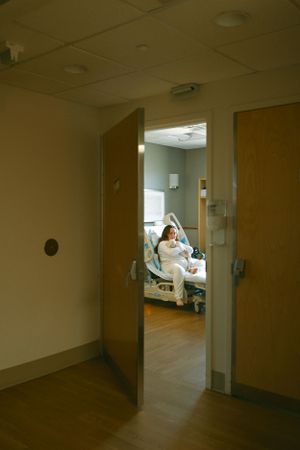 View through open hospital room doorway showing patient resting in adjustable bed.