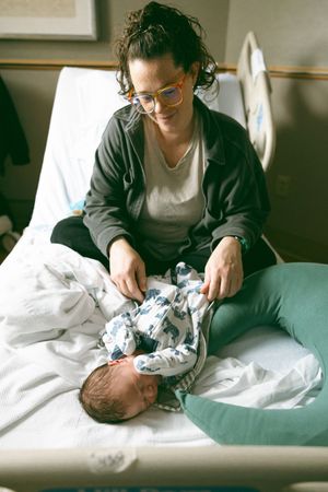 A person wearing glasses and a dark cardigan tends to a newborn baby on a white hospital bed.