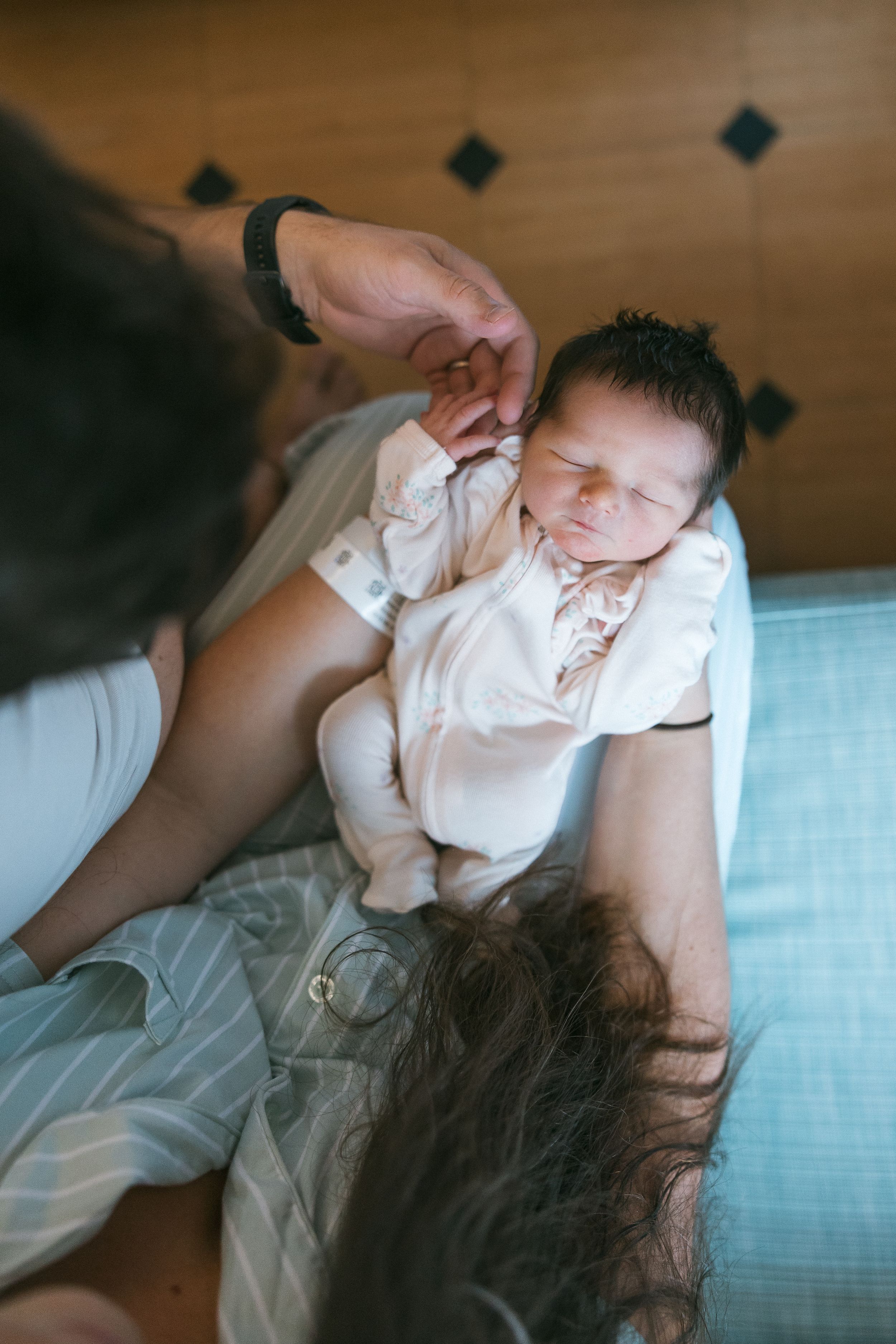 Newborn baby in white onesie cradled in parents arms during an intimate family moment at home.