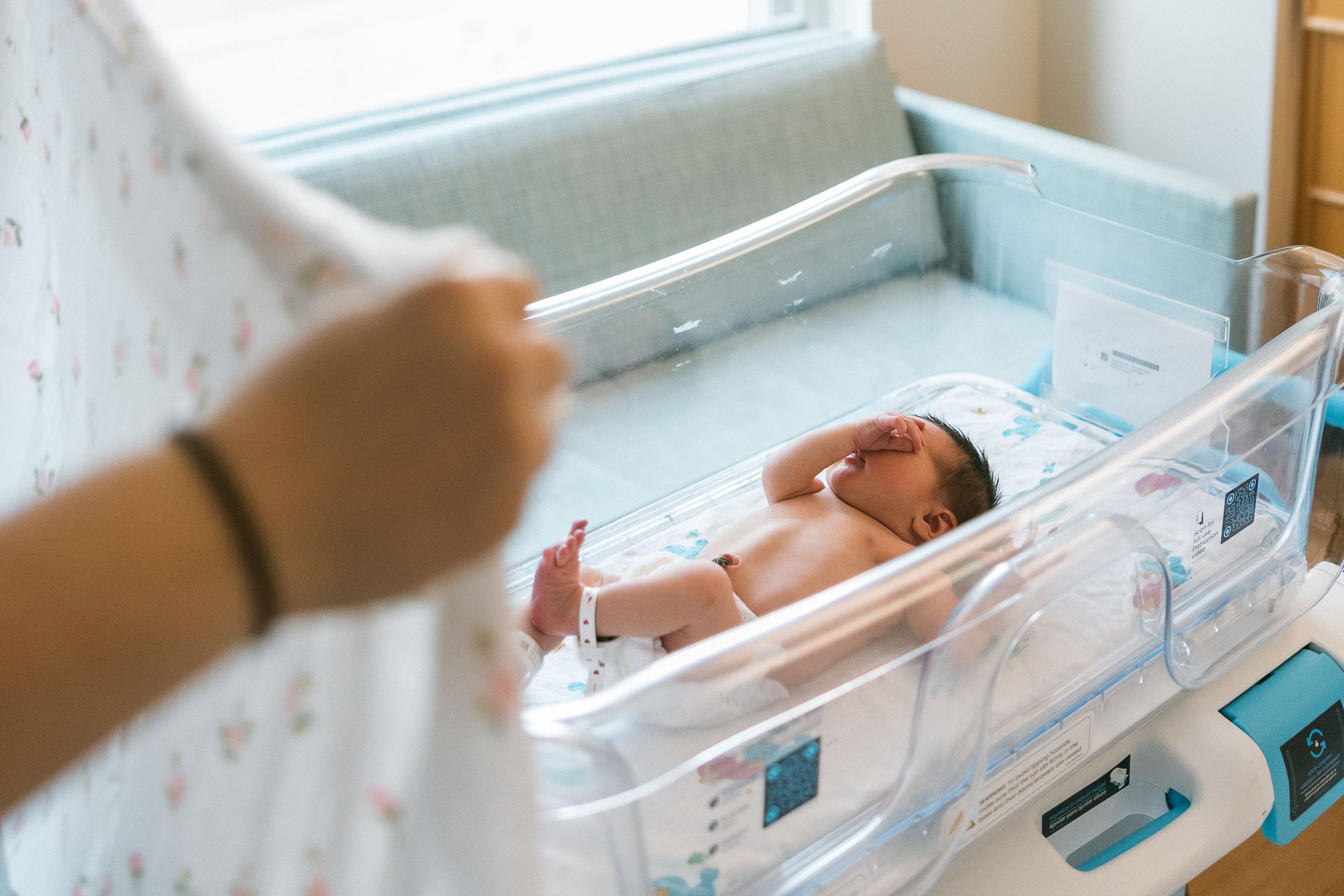 Newborn baby resting in a hospital bassinet while a parent reaches over to tend to the infant.