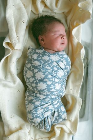 A newborn baby wrapped in a blue and white floral swaddle blanket rests on cream-colored fabric.