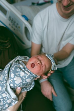 A baby wearing a floral patterned outfit smiles while being cradled by someone in a white shirt.