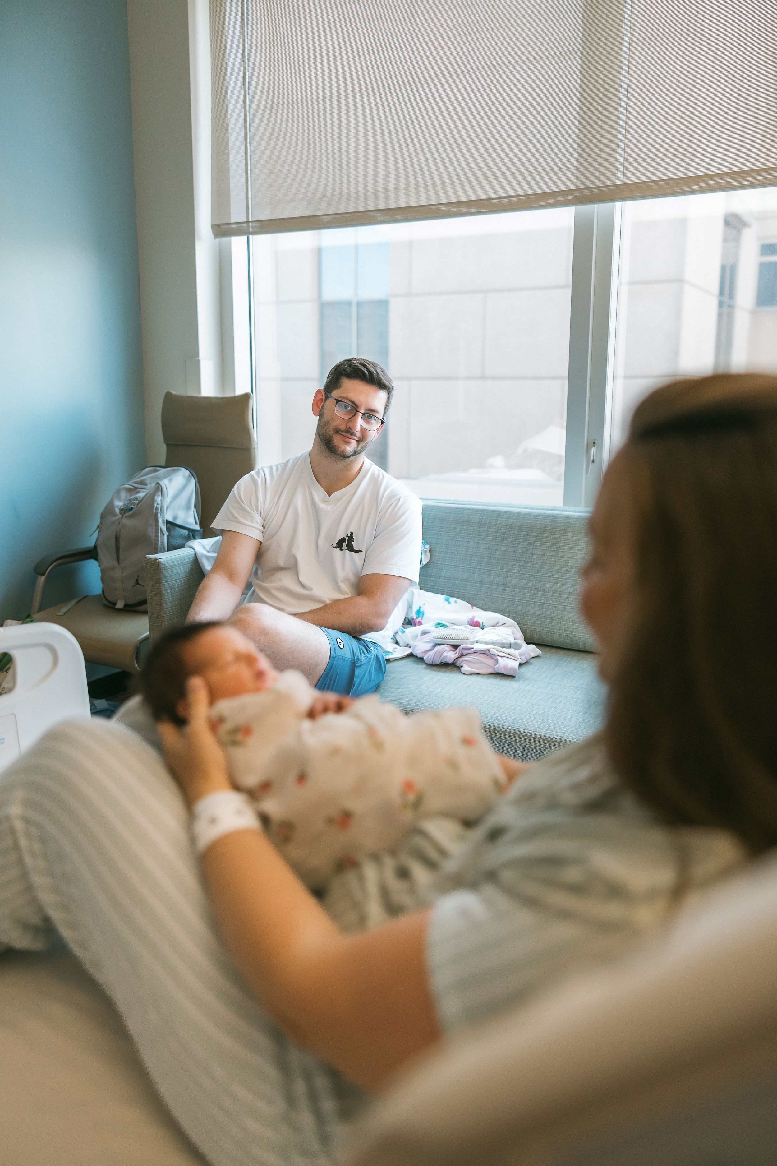 New parents share a tender moment in a hospital room, with dad smiling as mom holds their swaddled newborn baby.