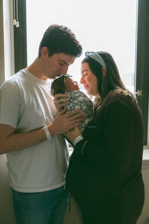 Parents sharing intimate moments with their newborn baby near a bright window.