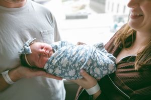 A newborn baby wearing a blue floral swaddle is cradled between two adults in a sunlit room.