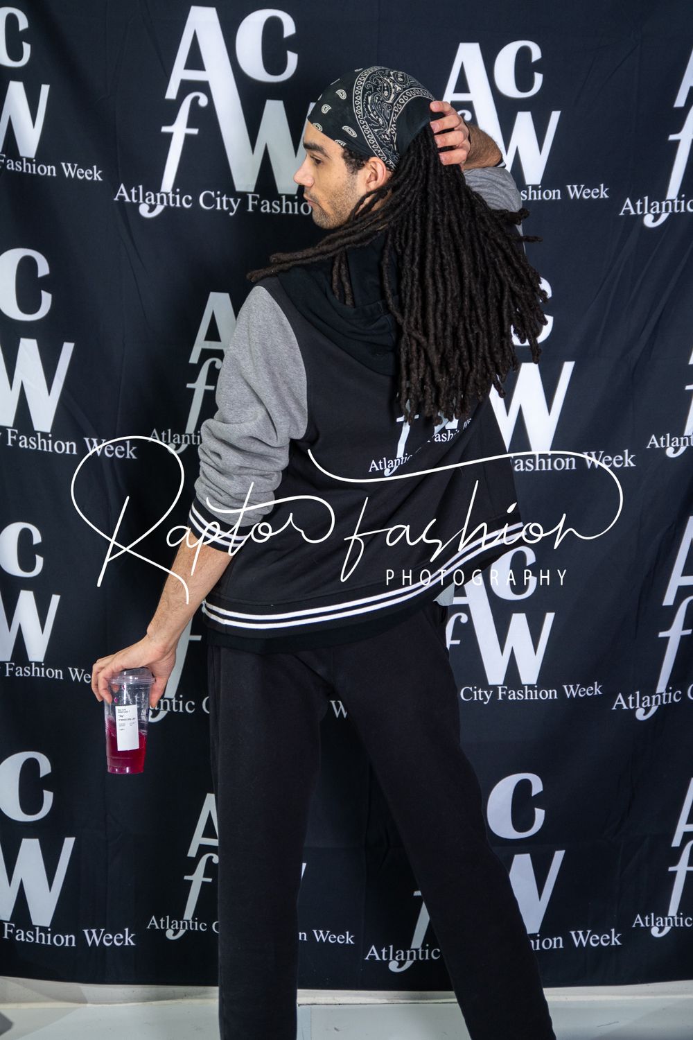 Attendee poses at fashion week event in front of ACW branded step-and-repeat backdrop while holding drink.