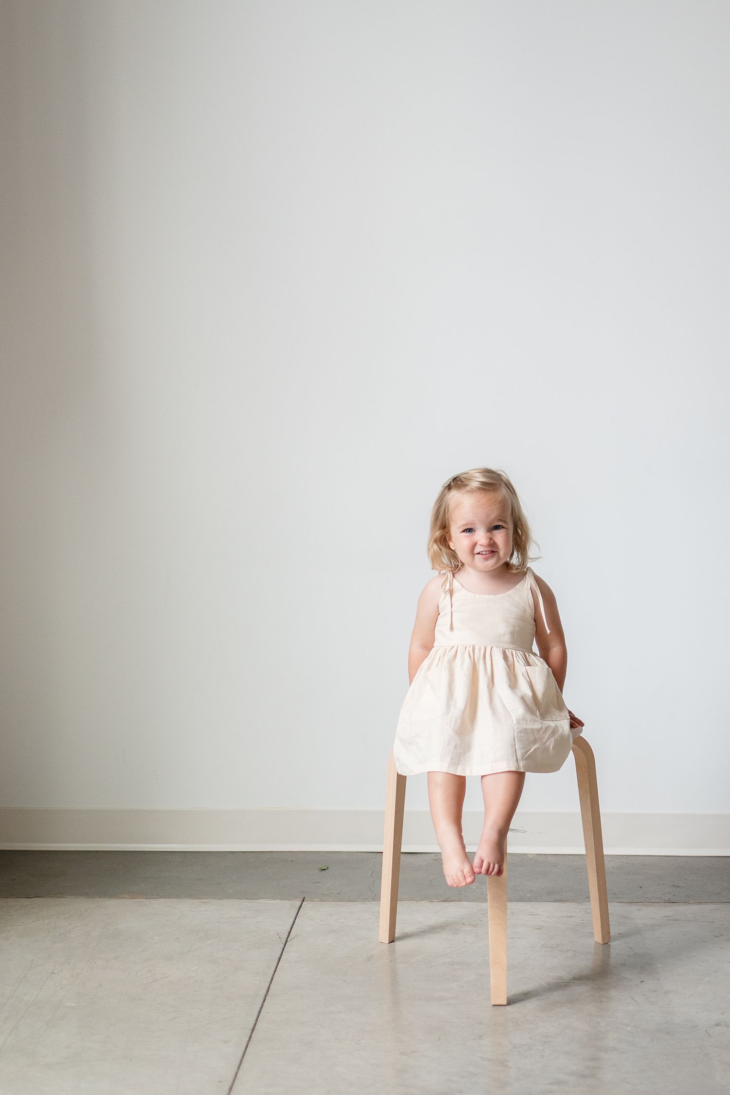 Little girl in a cream-colored sundress sits happily on a wooden stool against a minimalist white wall backdrop.