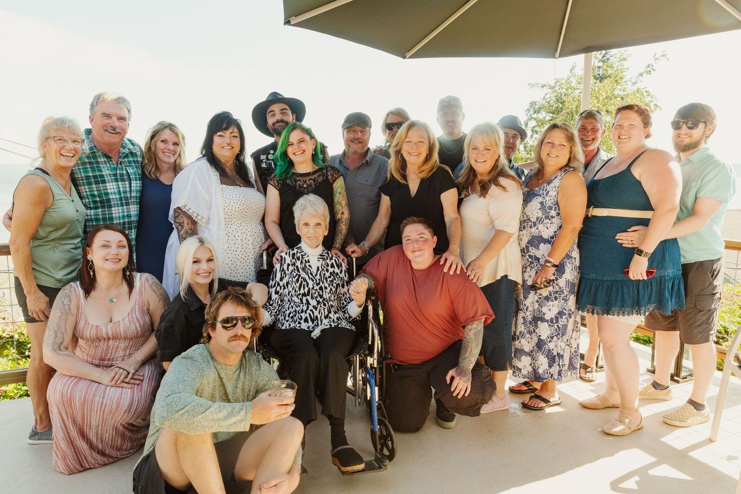 A diverse group of people gathered outdoors under a patio umbrella on a sunny day.