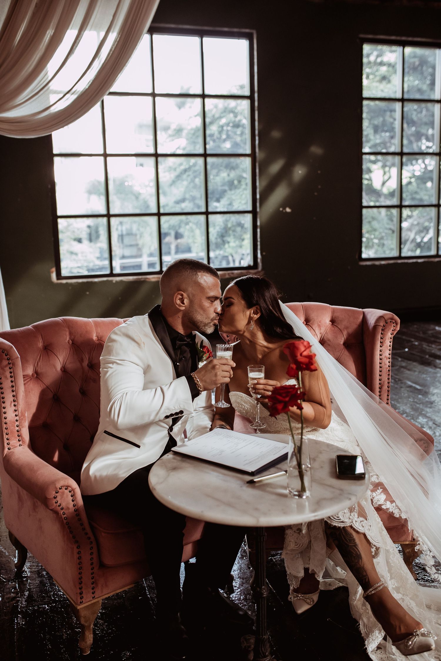 A romantic wedding couple shares an intimate moment on a pink velvet couch by large windows in a dark moody setting.