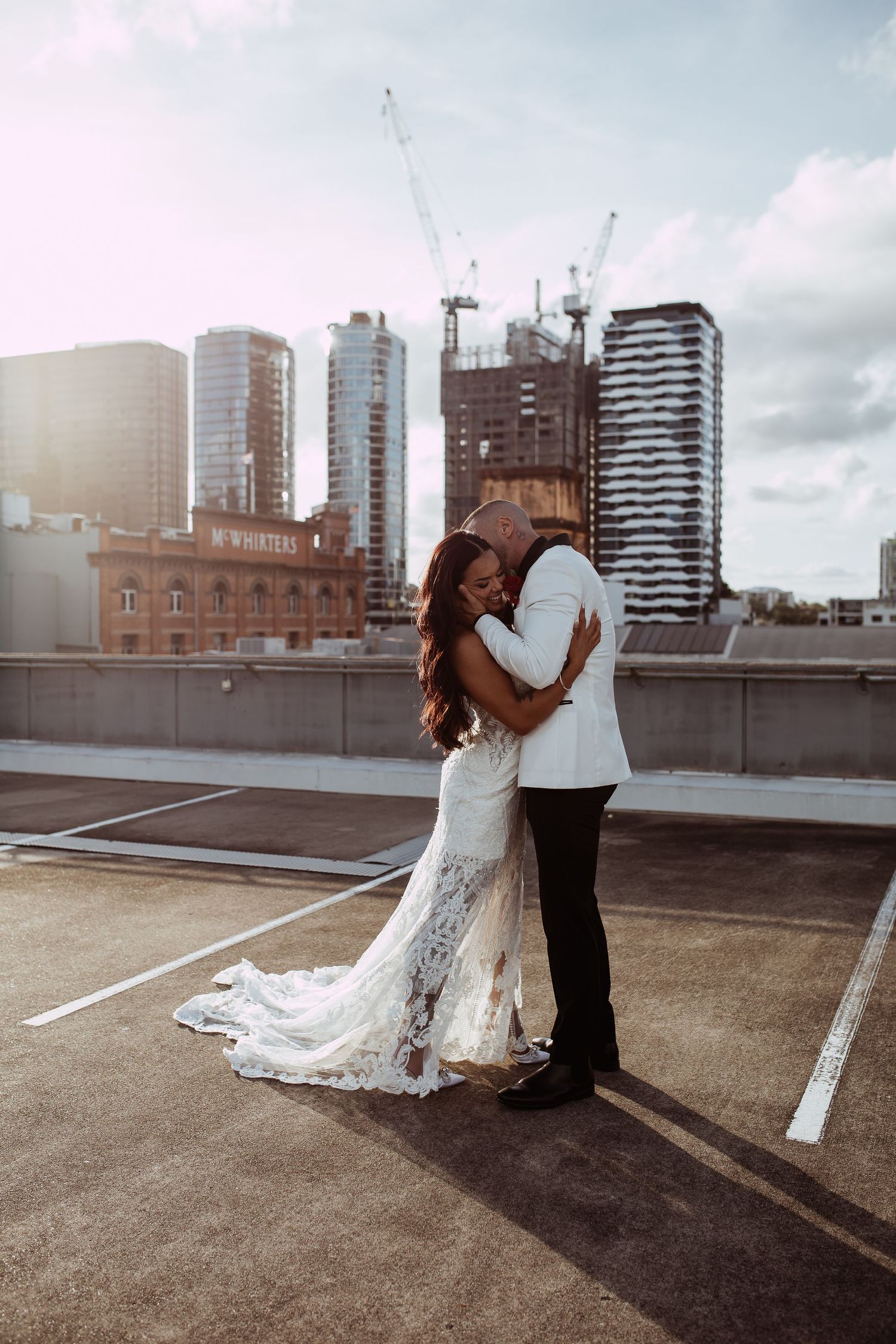 A romantic couple embrace on an urban rooftop parking lot with city skyscrapers and construction cranes in the background.