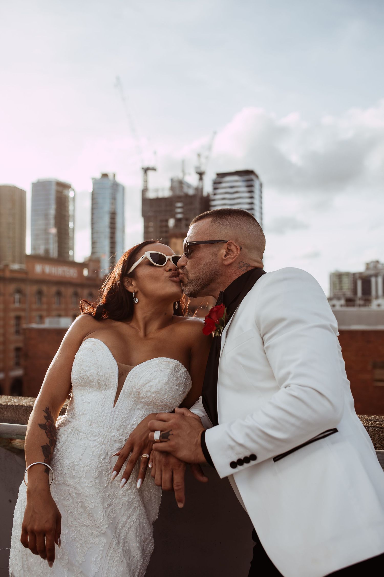 A newlywed couple shares a romantic kiss on a rooftop with city buildings in the background at sunset.