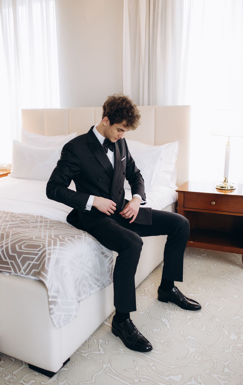 Young man in a tailored black suit and tie adjusts his jacket while seated on a white bed in an elegant room.