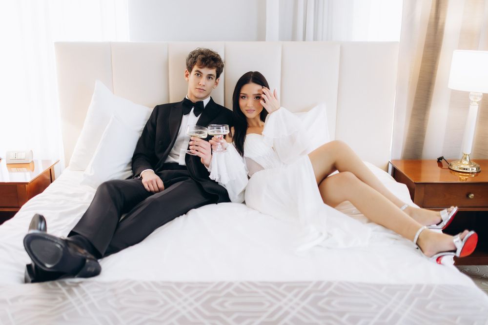 A couple in wedding attire relaxes on a white hotel bed with wine glasses while wearing stylish formal wear.
