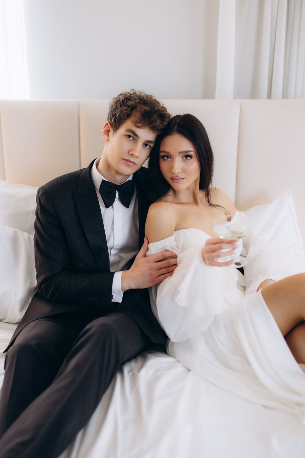 An elegant wedding couple in formal attire poses intimately on a white sofa against light curtains.