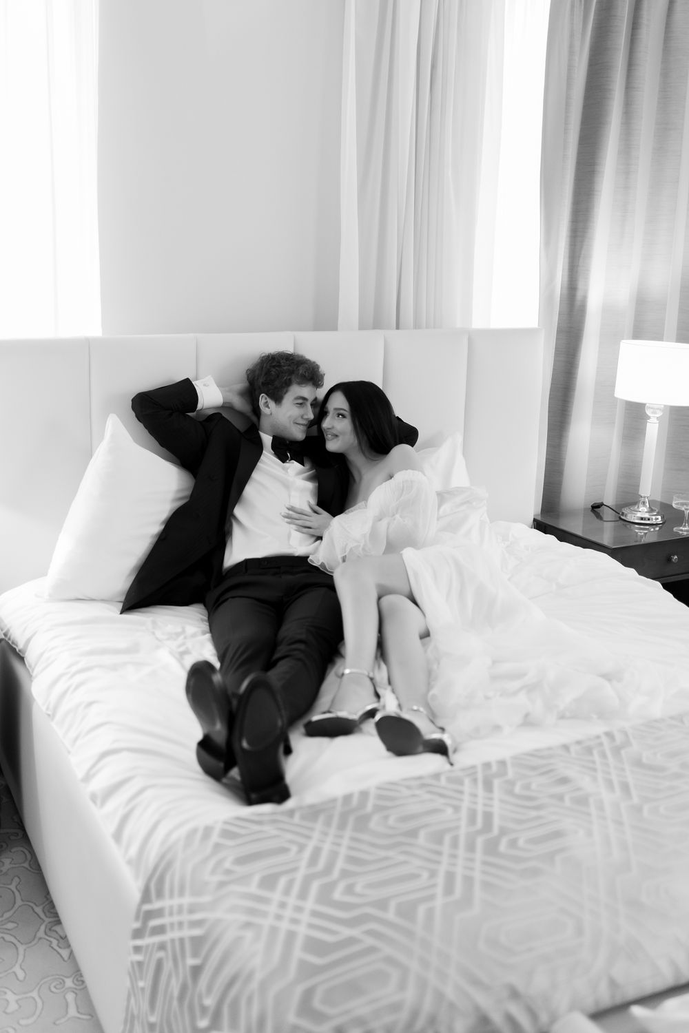 Black and white romantic photo of a couple in formal attire lying on a bed with white bedding in an elegant hotel room.
