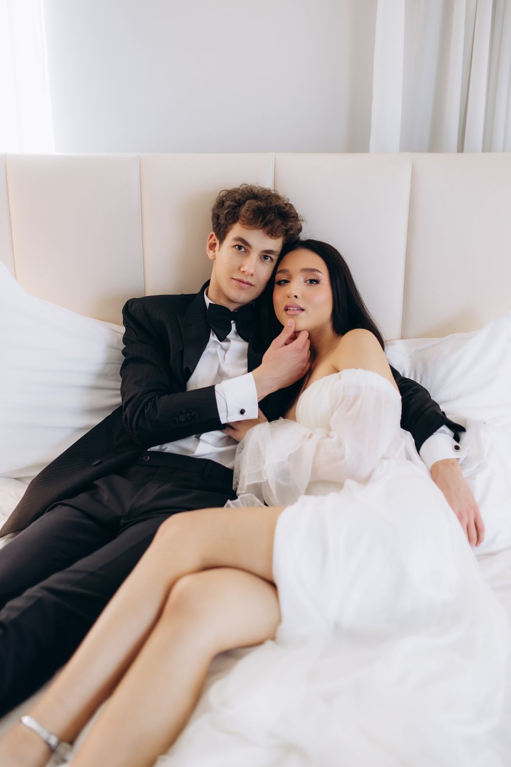 A bride in white dress and groom in black tuxedo pose intimately on white couch during wedding photoshoot.