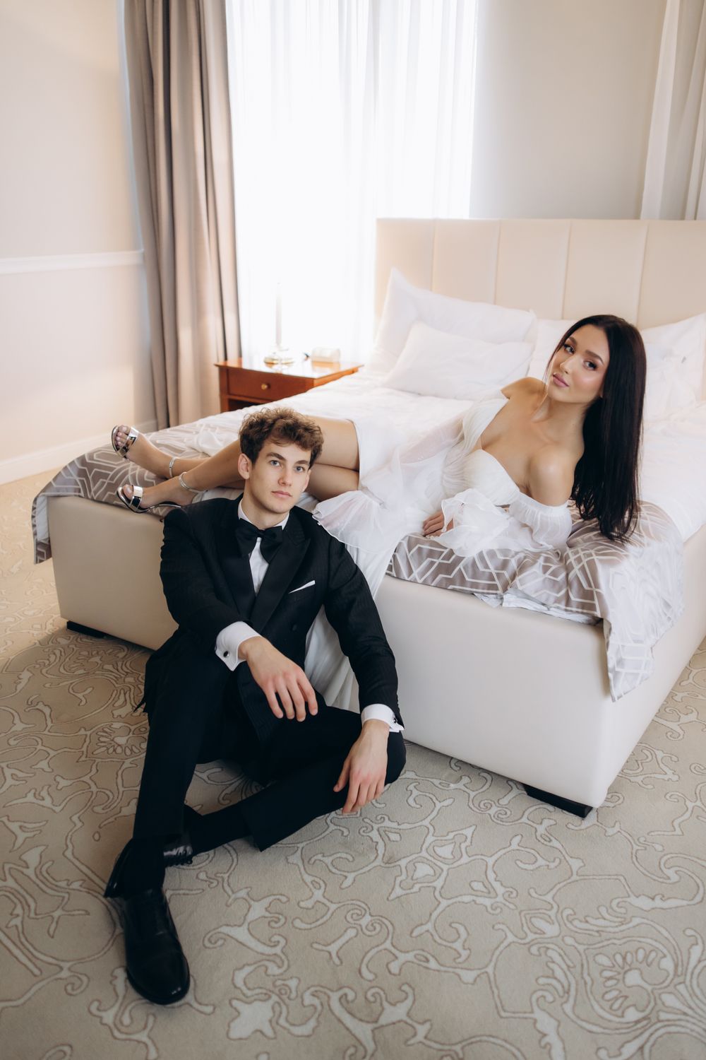 Couple in black tuxedo and white dress lounging elegantly in a contemporary room setting with patterned carpet.