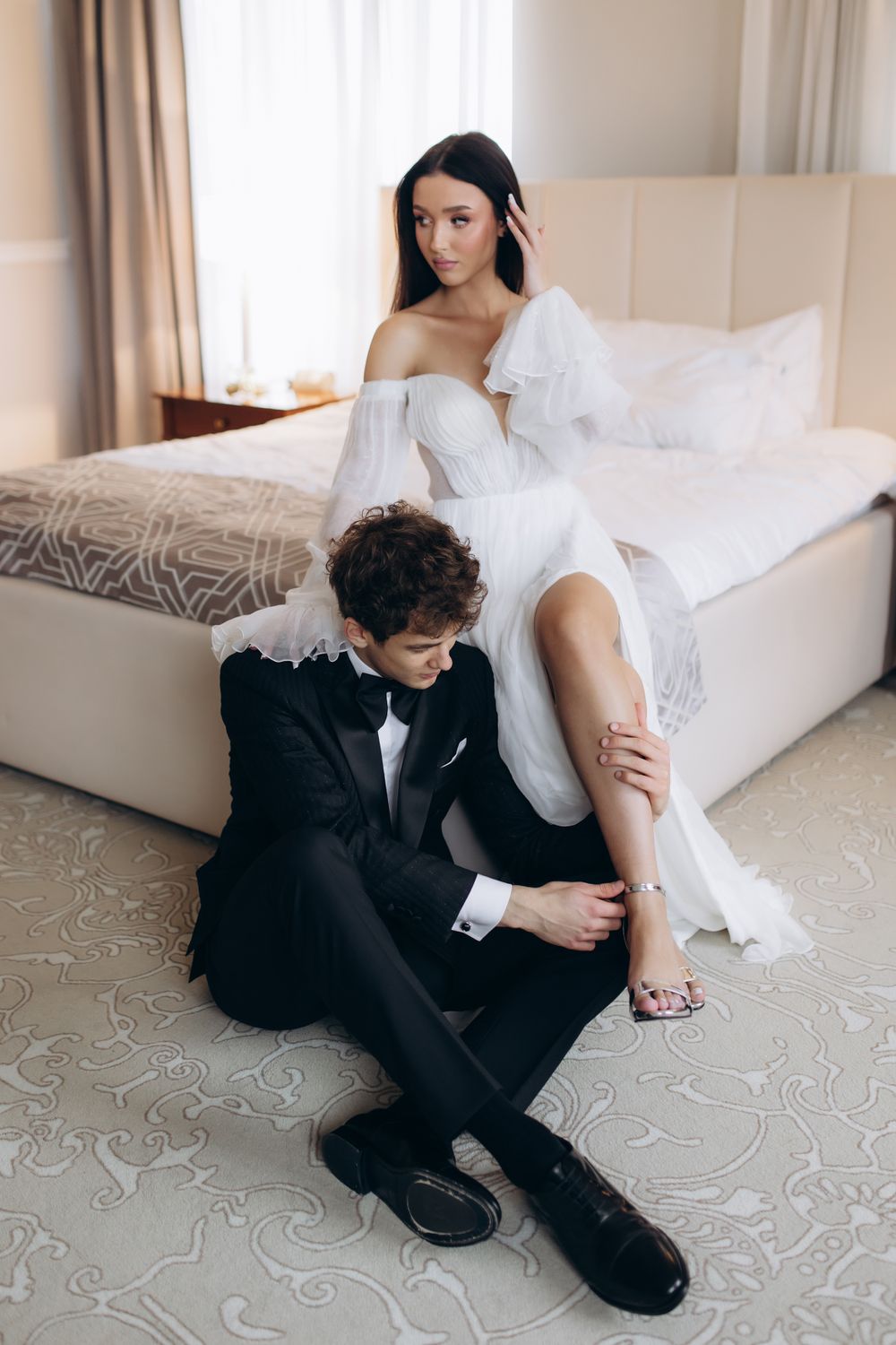 A newlywed couple in formal wedding attire sharing a tender moment in an elegant hotel room with a white bed.